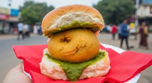 A close-up of a vada pav, a popular Indian street food, held in a hand. The vada pav consists of a deep-fried potato fritter (vada) sandwiched in a soft bread roll (pav), generously slathered with green chutney. The background is blurred, showing a street scene.